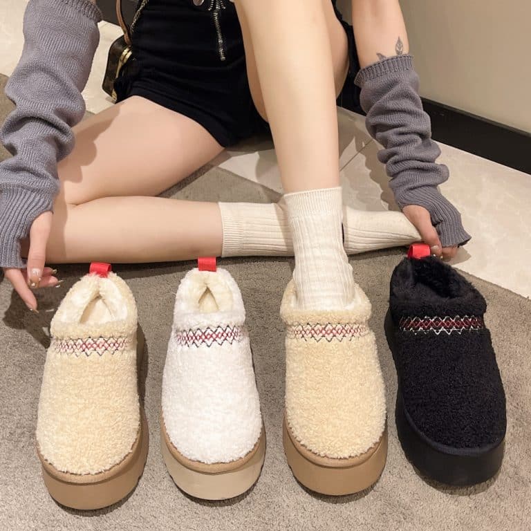Woman sitting on the floor wearing beige sheep wool slippers, next to a black pair.