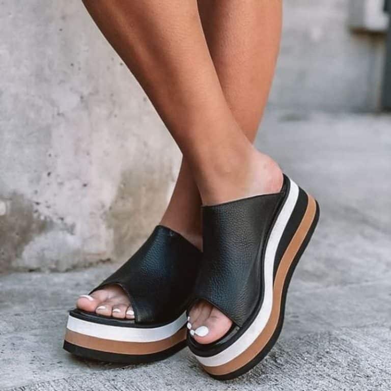 Orthopedic black leather slides for women, with striped platform sole, worn against a concrete wall.